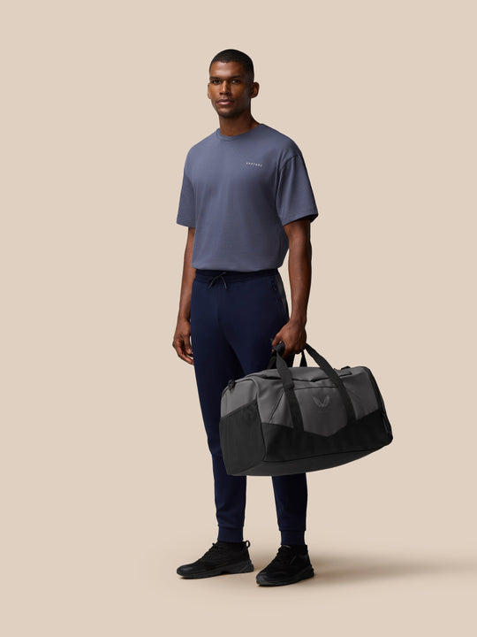 Male model holds a grey and black Core Holdall on a beige background