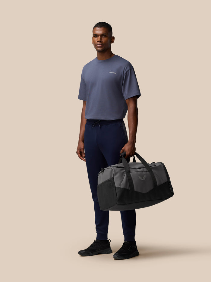 Male model holds a grey and black Core Holdall on a beige background