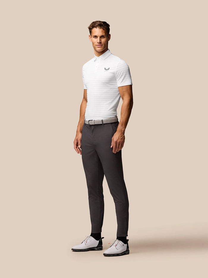 Full-body shot of a man in a white golf polo with Castore logo on the chest, paired with gray trousers.