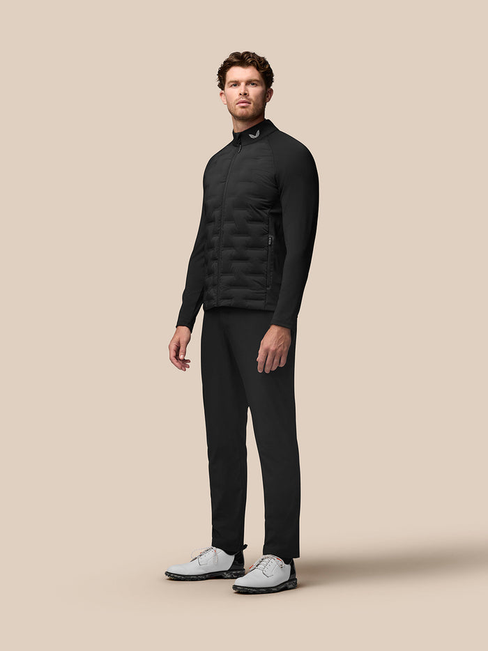 Full body view of a man in black Pinnacle Hybrid Golf Jacket with zip, standing against beige backdrop