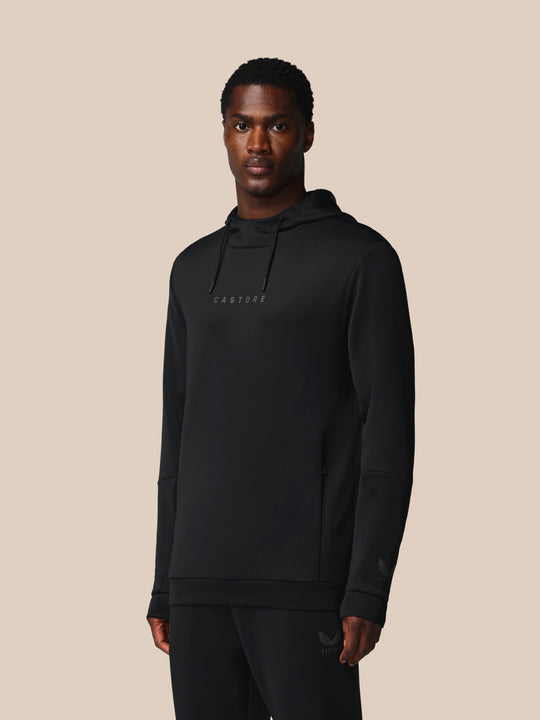 Male model wearing black Castore Flex Long Sleeve Hoodie with hood and Castore logo on the chest against beige backdrop.