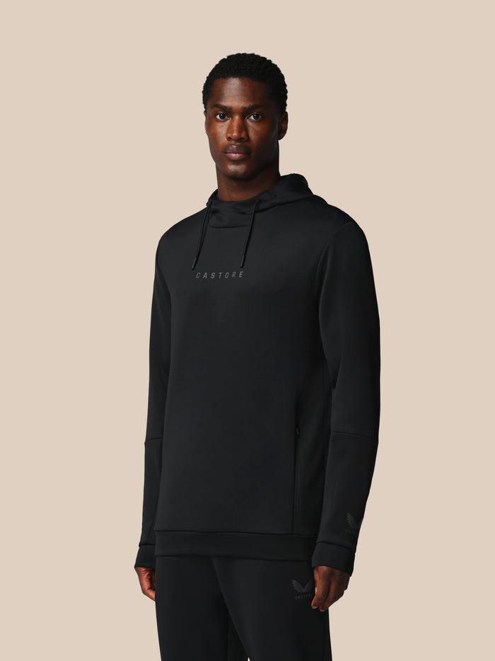 Male model wearing black Castore Flex Long Sleeve Hoodie with hood and Castore logo on the chest against beige backdrop.