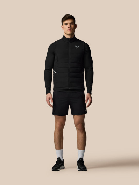 Volledig beeld van een man in een zwarte Apex Training Gilet over een mouwloze basislaag, tegen beige achtergrond