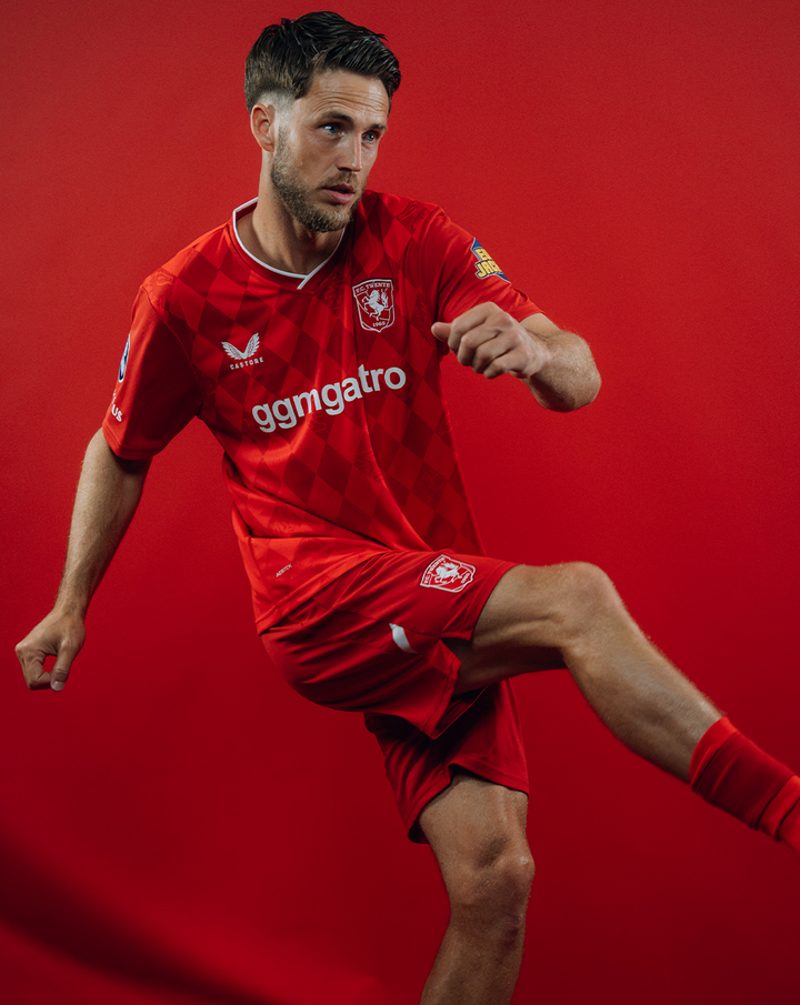 Speler in rode FC Twente voetbaloutfit in actie tegen rode achtergrond