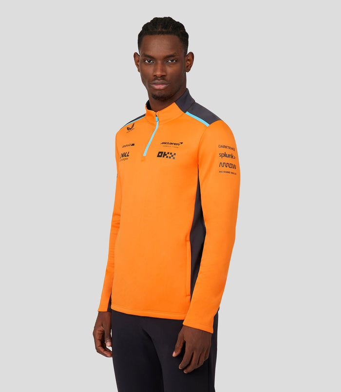 Male model wearing bright orange McLaren replica quarter zip top with blue zipper and dark shoulder panels, featuring printed McLaren Formula 1 Team logo and sponsor logos.