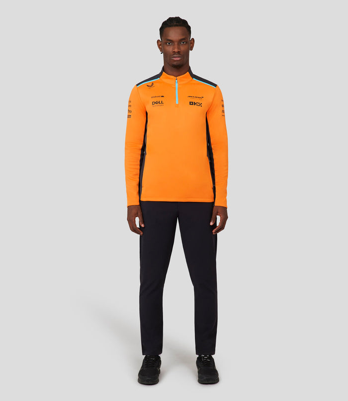 Full front view of a male model wearing a bright orange McLaren replica quarter zip top with sponsor logos and a blue zipper.